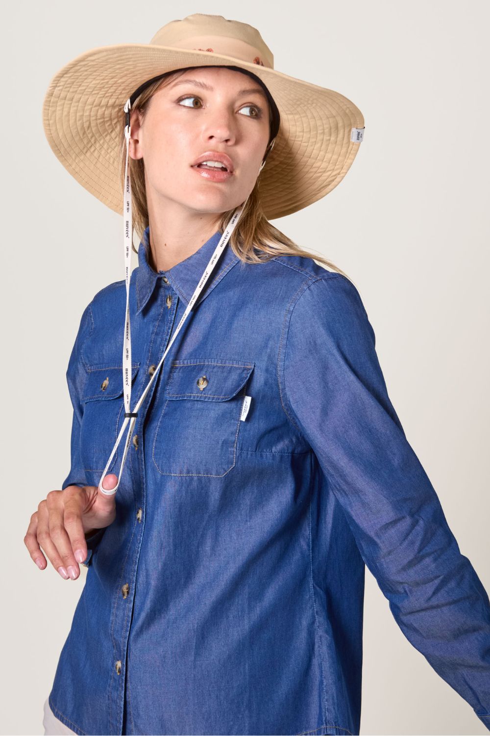 Chapeau anti-UV beige UPF50+ porté par une femme en chemise bleue, idéal pour la protection solaire en extérieur.